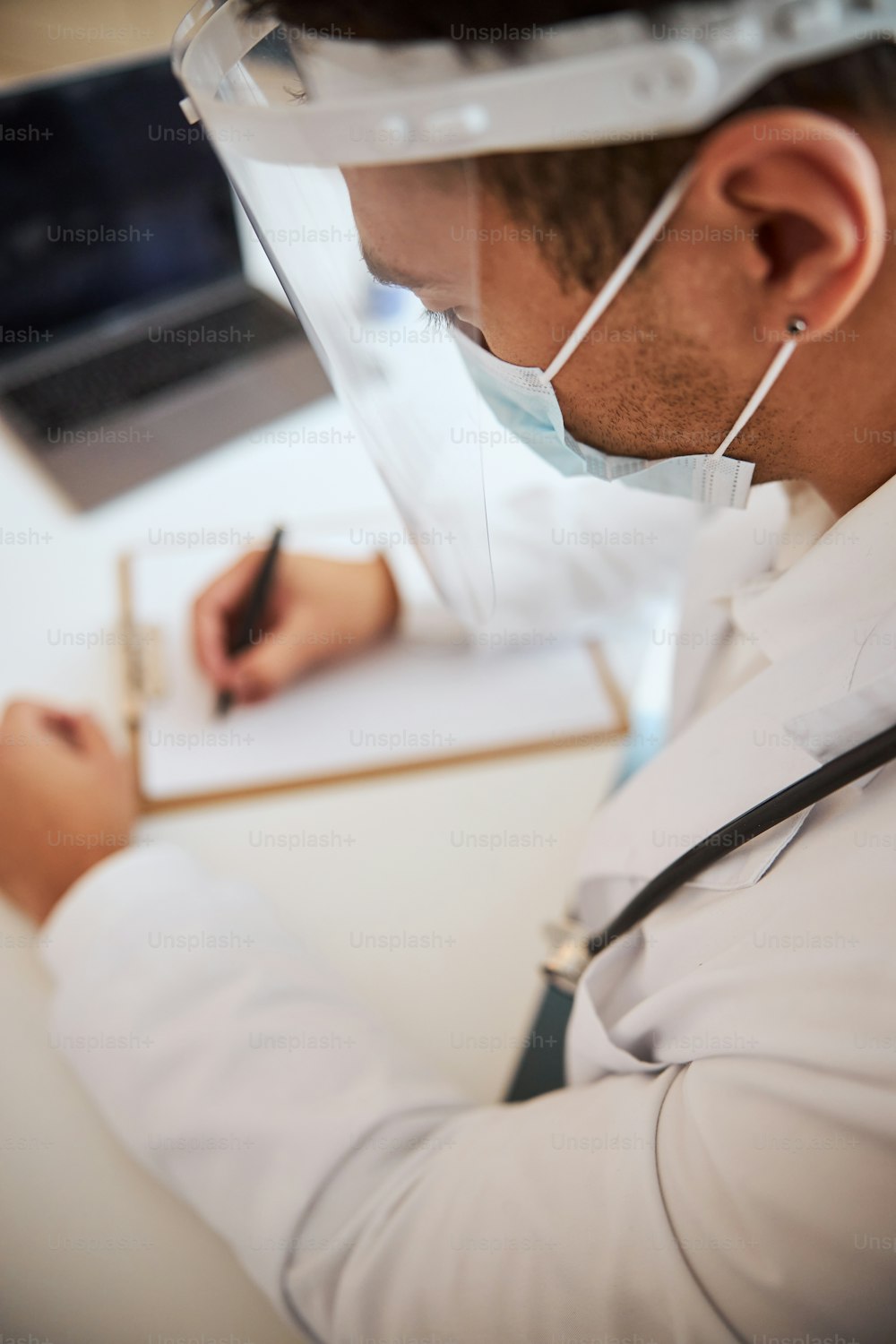 Abgeschnittenes Kopfporträt eines unbekannten Arztes in weißer Uniform, der am Schreibtisch sitzt und etwas in die Zwischenablage im Schrank schreibt
