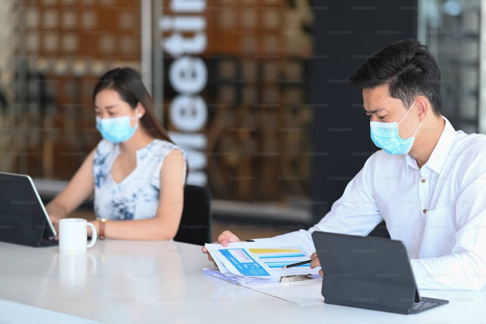 Two young business people in protective mask working together with modern device in office.