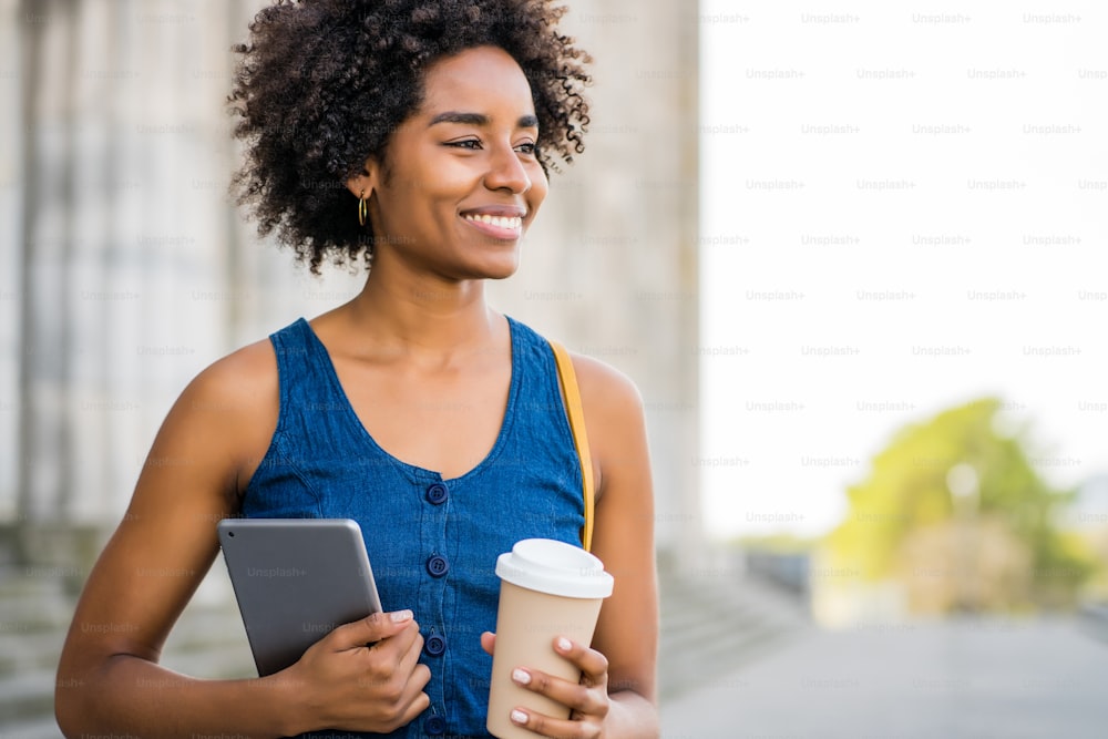 Portrait of afro business woman holding a digital tablet and a cup of coffee while standing outdoors on the street. Business and urban concept.