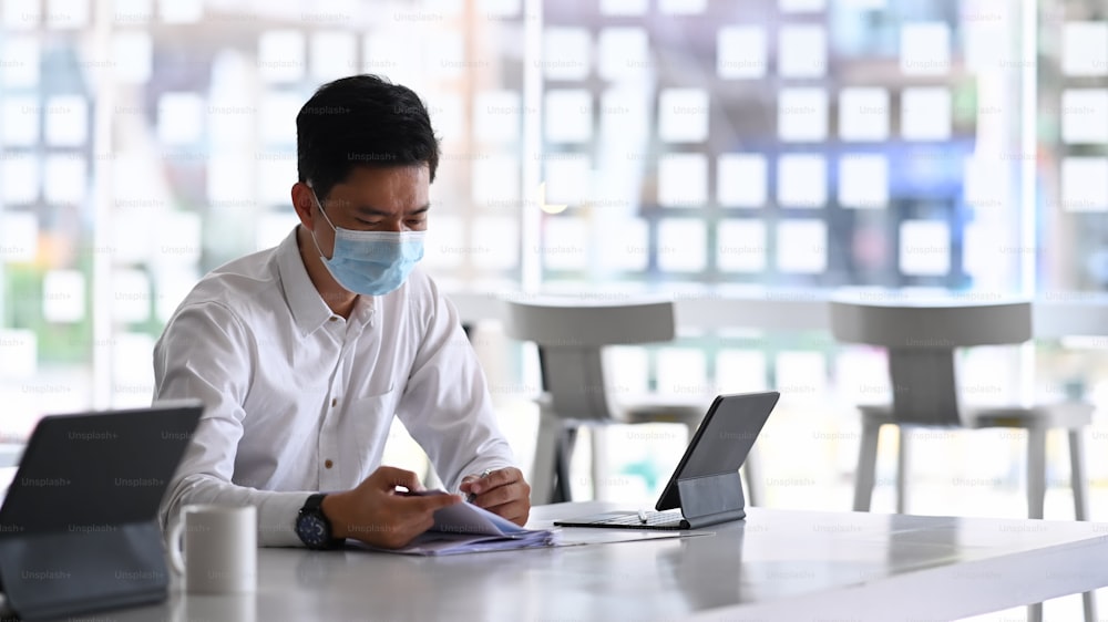 Businessman wearing protective mask working with tablet computer and analyzing business data on office desk.