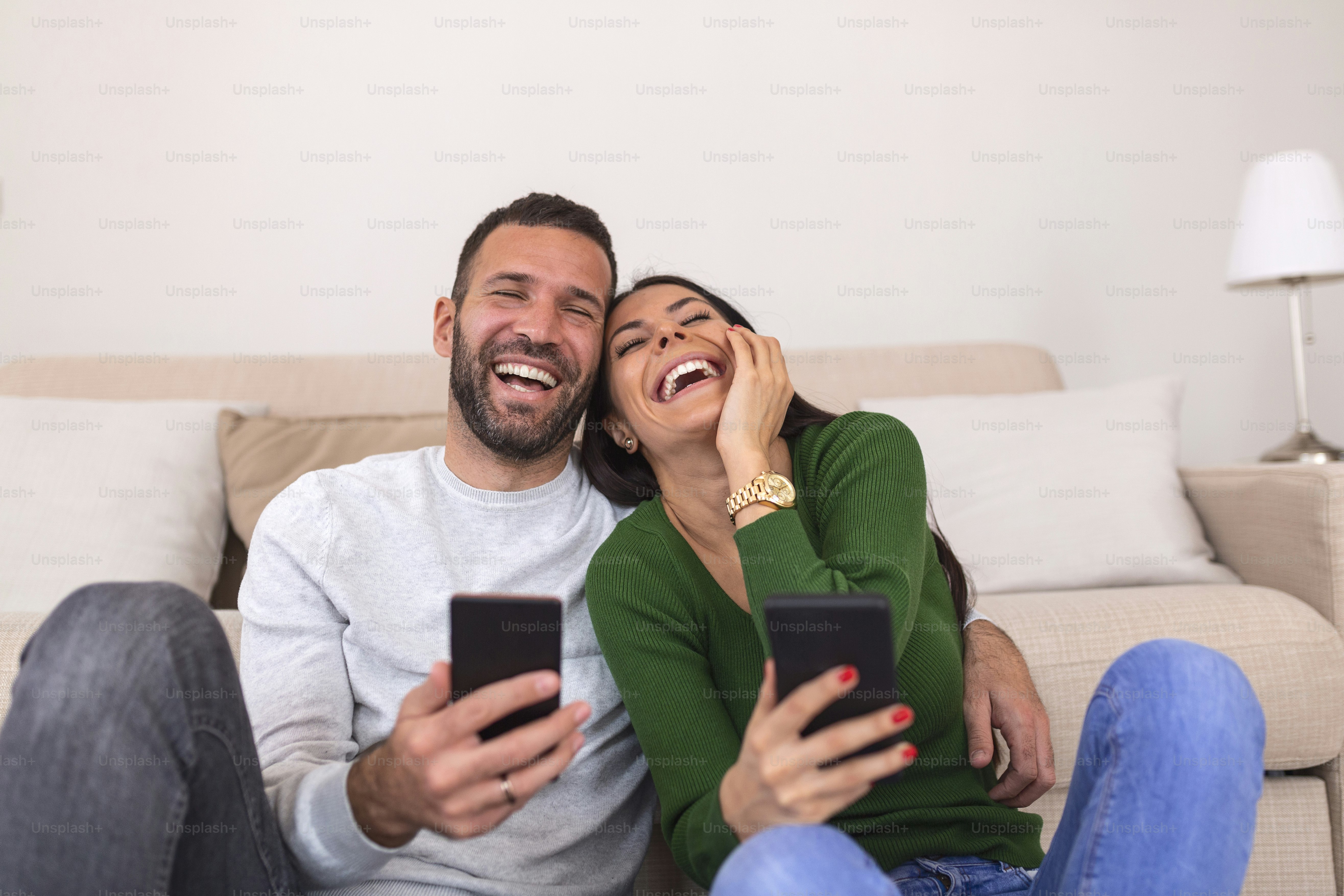 Happy young wife showing funny video in social network on cellphone to laughing husband. Smiling spouses shopping online web surfing, relaxing together on sofa in modern studio kitchen living room.