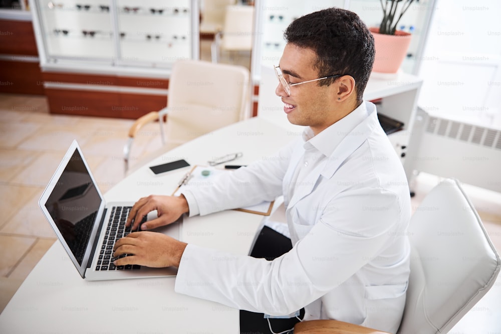 Oftalmólogo sonriente, satisfecho, con gafas, con bata de laboratorio, sentado frente a la computadora portátil en su oficina
