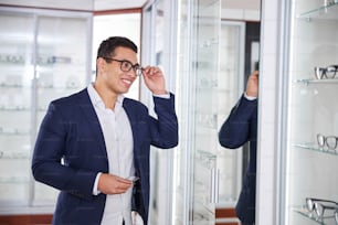 Attractive joyful young dark-haired Latin American male client trying on eyewear in an optical store