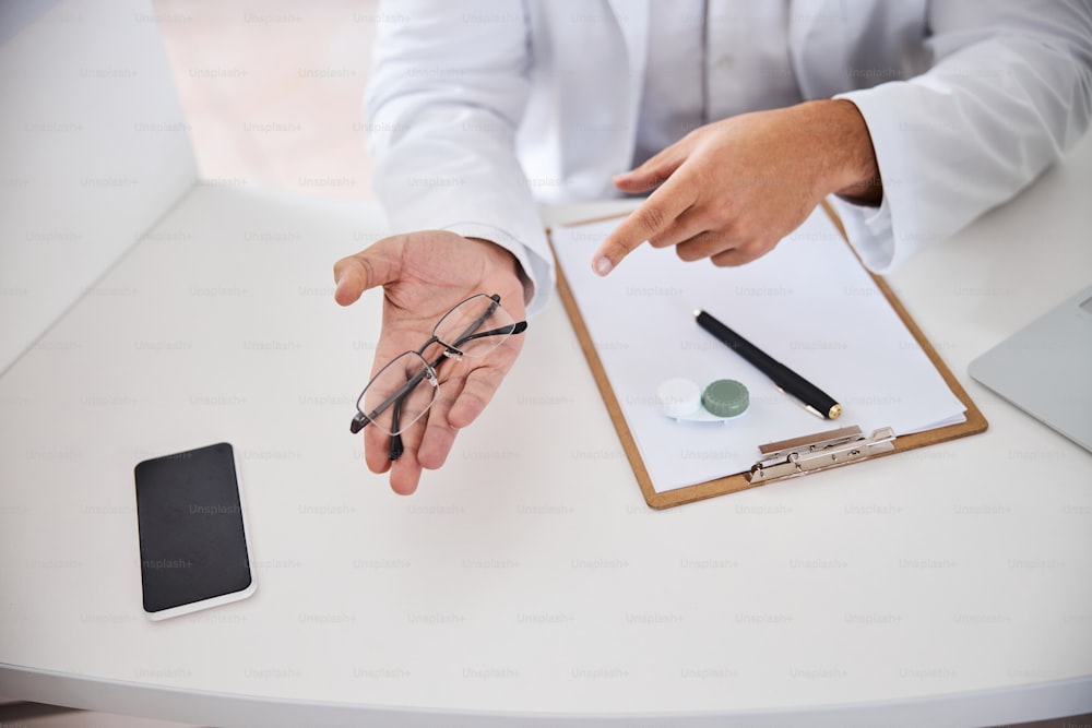 Cropped photo of an optician in a lab coat pointing with his forefinger at spectacles on his palm