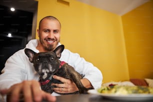 Smiling pleased male in a terry bathrobe and his calm canine friend sitting at the table in a hotel room
