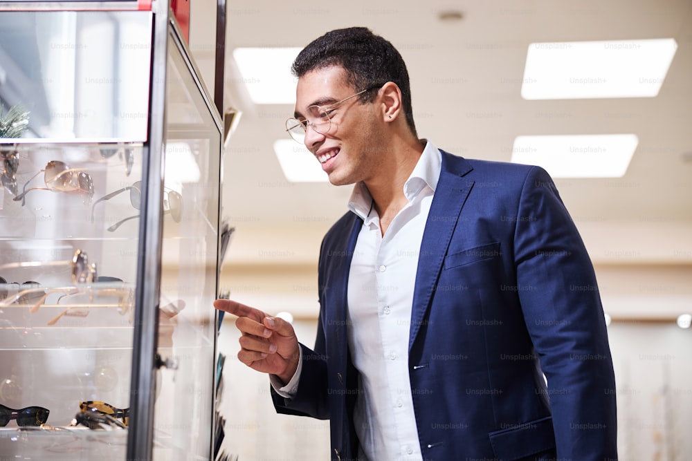 Joyous young male buyer pointing with his index finger at the eyewear behind the glass