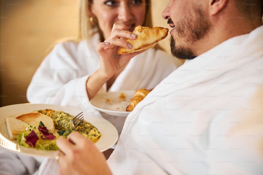 Photo recadrée d’un homme caucasien souriant avec une assiette de nourriture mangeant un croissant de la main féminine