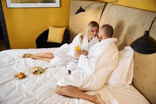 Beautiful blonde lady and her young male spouse in terry bathrobes clinking orange juice glasses