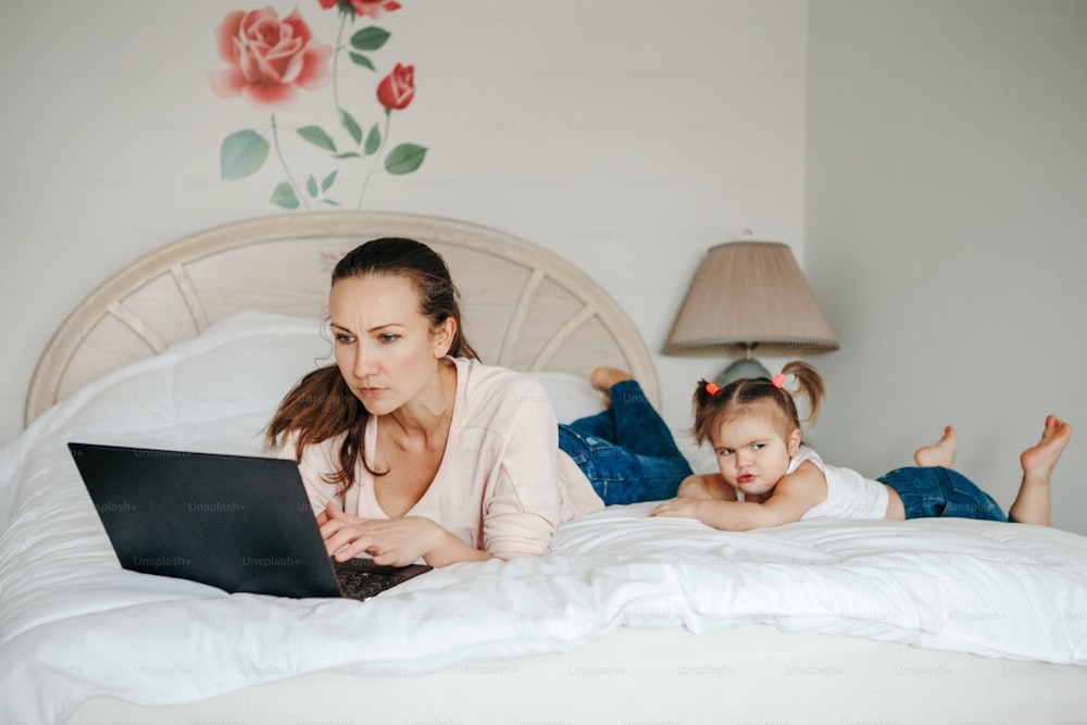 子供、子供、子供と一緒に在宅勤務。寝室でノートパソコンで作業する母親と、傍らに幼い娘の幼児がいる。面白い率直な家族の瞬間。コロナウイルス検疫ロックダウン中のニューノーマル。