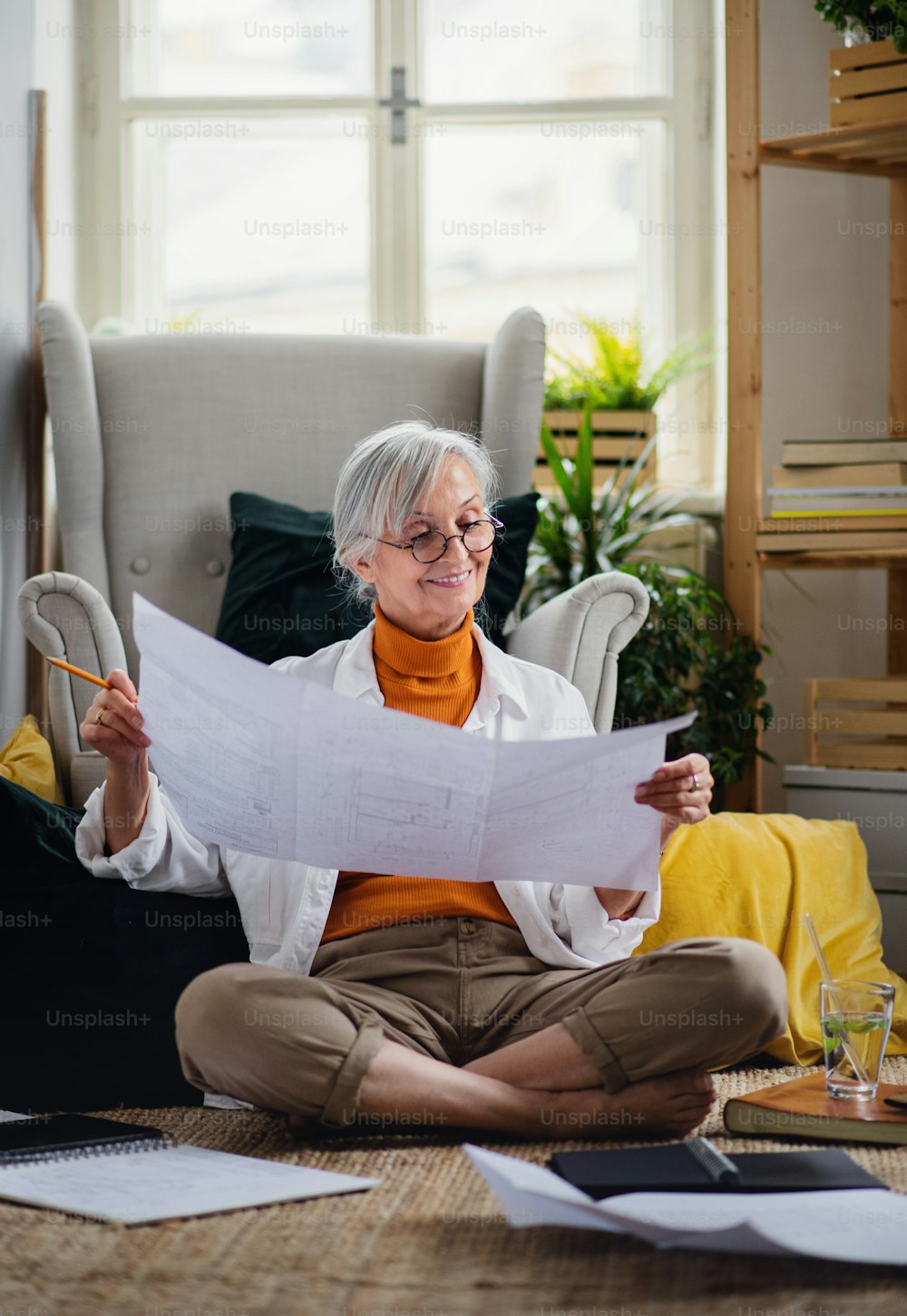 Portrait of senior woman architect sitting with blueprints on floor indoors at home, working.