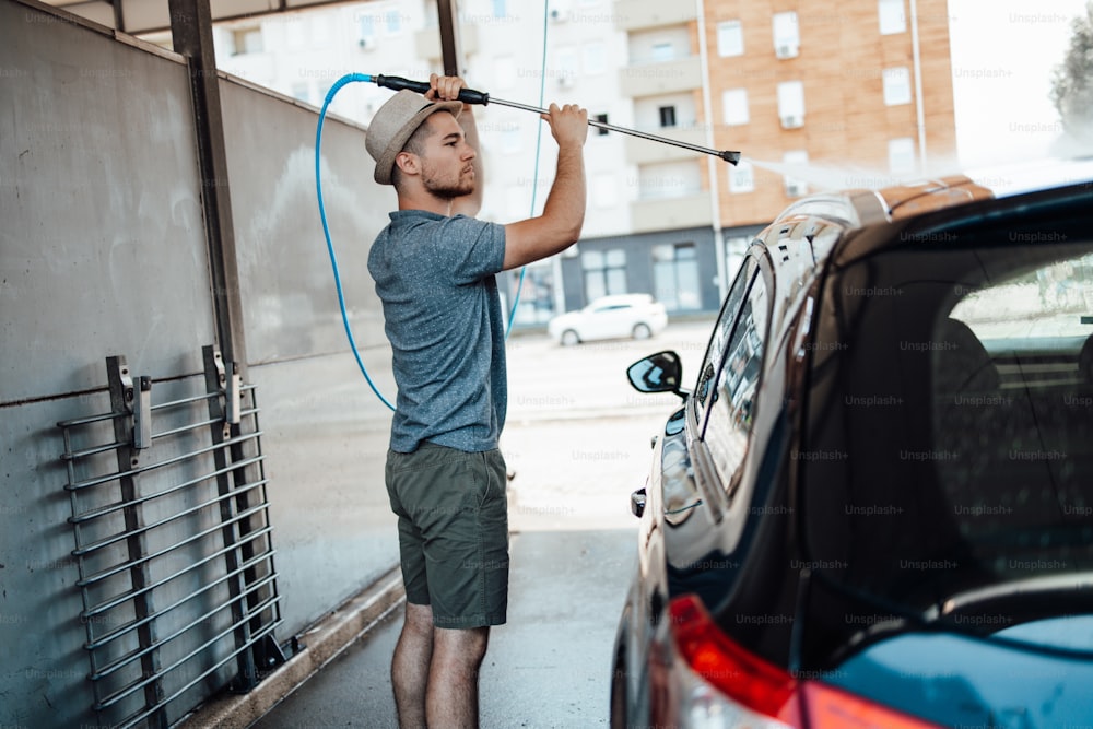 Joven con sombrero lavando su auto durante el día en la estación de lavado de autos usando agua a alta presión.