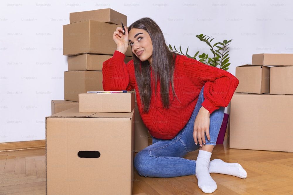 Una joven hermosa mujer hace sus notas y planes en su nuevo piso, apartamento. Mujer joven empacando caja de cartón. Concepto de casa de mudanza.