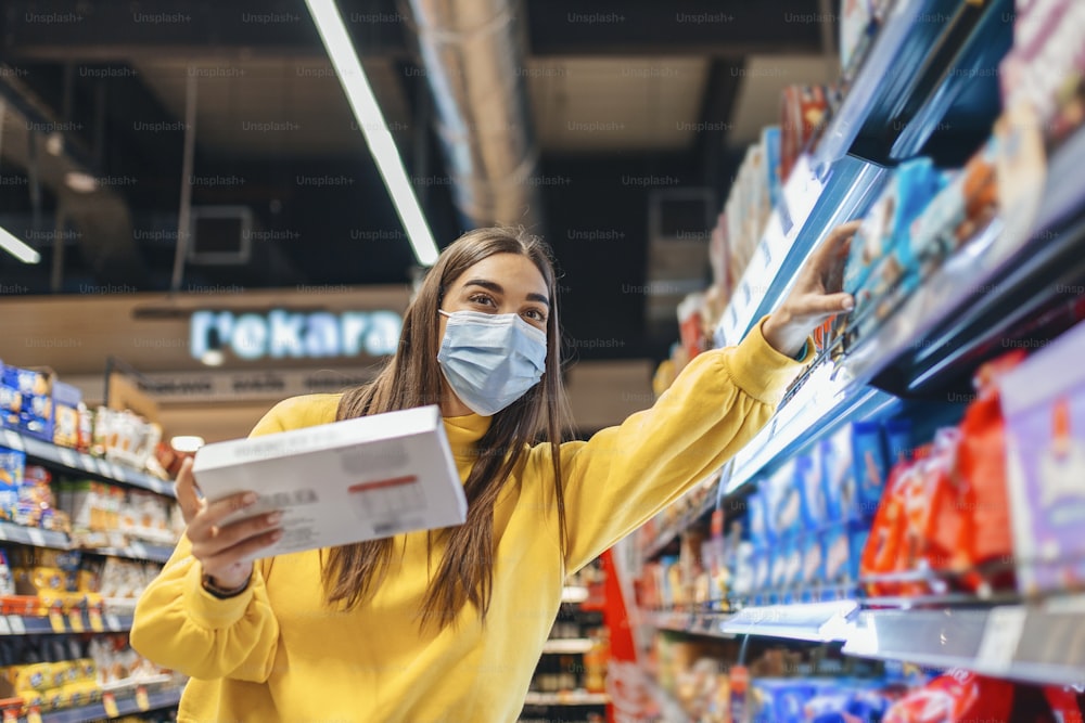 Distanziamento sociale in un supermercato. Una giovane donna con una maschera facciale usa e getta che compra cibo e lo mette in un cestino della spesa. Fare shopping durante l'epidemia di Coronavirus Covid-19