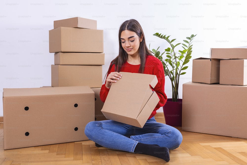 Mujer joven empacando cajas, mudándose a un nuevo apartamento. Mujer joven que mueve nuevo lugar y concepto de reparación - mujer joven feliz con muchas cajas de cartón que se sientan en el piso