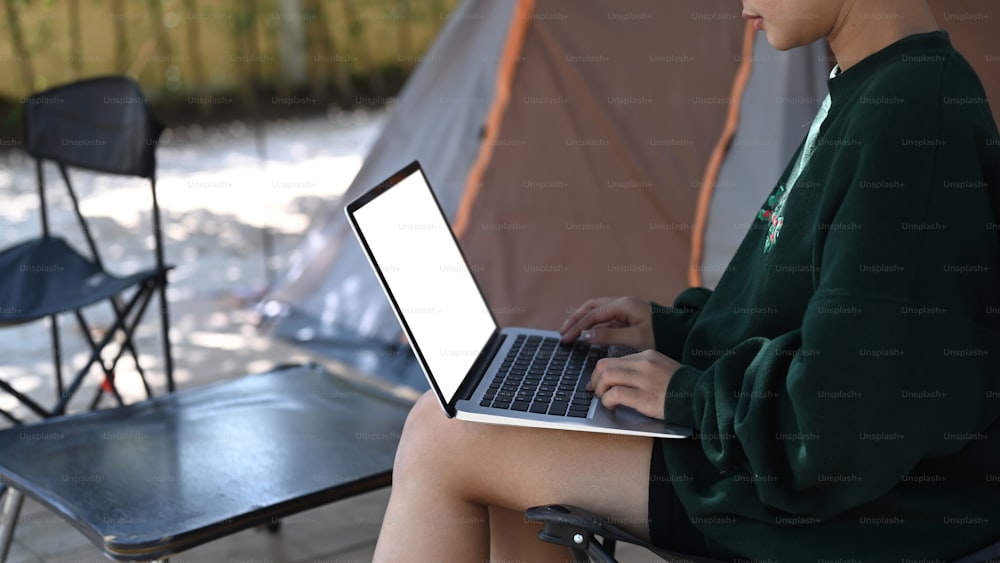 Vista lateral de una joven asiática que usa una computadora portátil mientras está sentada cerca de la tienda durante el campamento en el campo.
