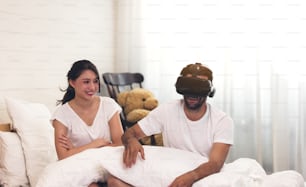 Woman in bed her husband man with short dark hair and using his VR head set gaming video with the 3D glasses, having fun.