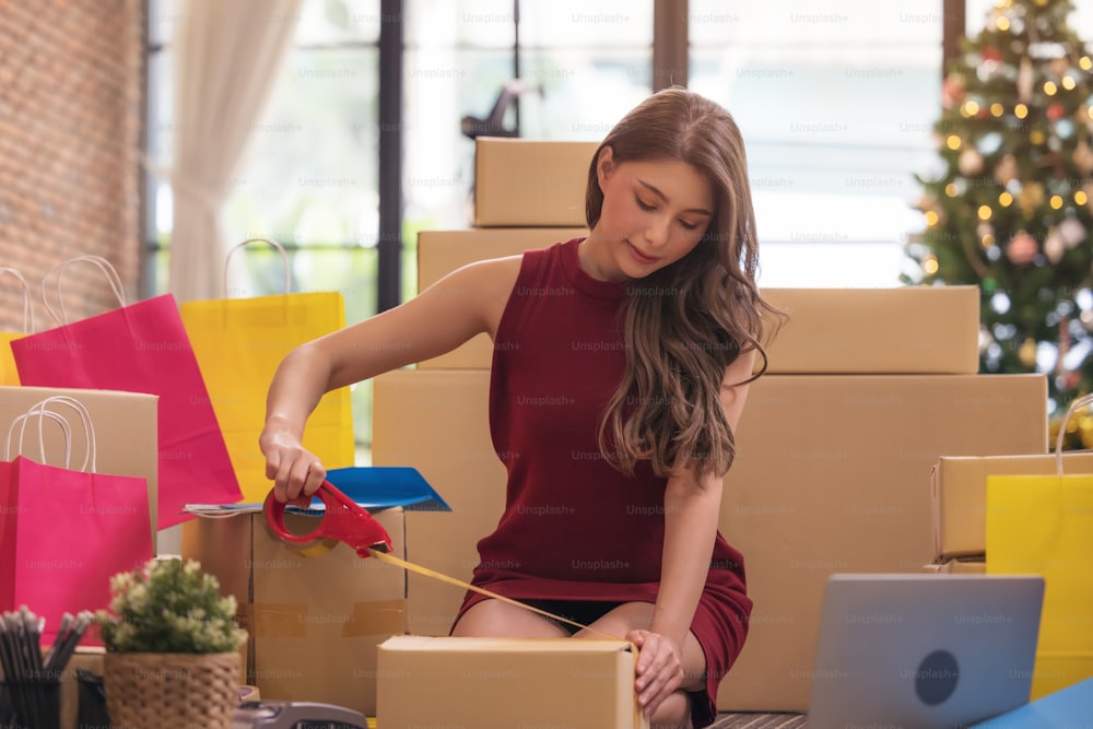 Femme d’affaires scotchant une boîte en carton, femme asiatique emballant des boîtes parmi une pile de colis dans son entreprise d’achat en ligne à la maison