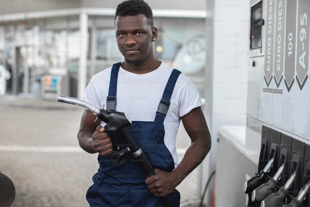 Handsome young African guy, gas station worker, wearing white t-shirt and blue overalls and holding filling gun. Close up front view.