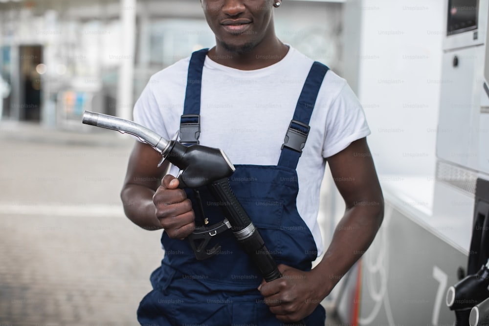 作業服を着た黒い肌の男、ガソリンスタンドの従業員、ガソリンスタンドの屋外に立って、燃料銃のノズルを手に持っているトリミングされた画像の接写。