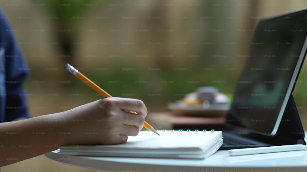 Vista lateral de la mano que sostiene el lápiz para escribir en un cuaderno en blanco mientras estudia en línea en el jardín de su casa