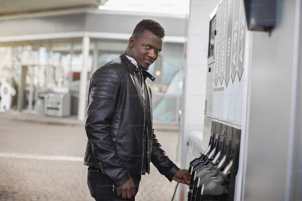 Junger lächelnder, angenehmer afrikanischer Mann, der vor der Kamera posiert, während er an der Tankstelle steht, bereit, sein Luxusauto mit der Tankpistole zu betanken.