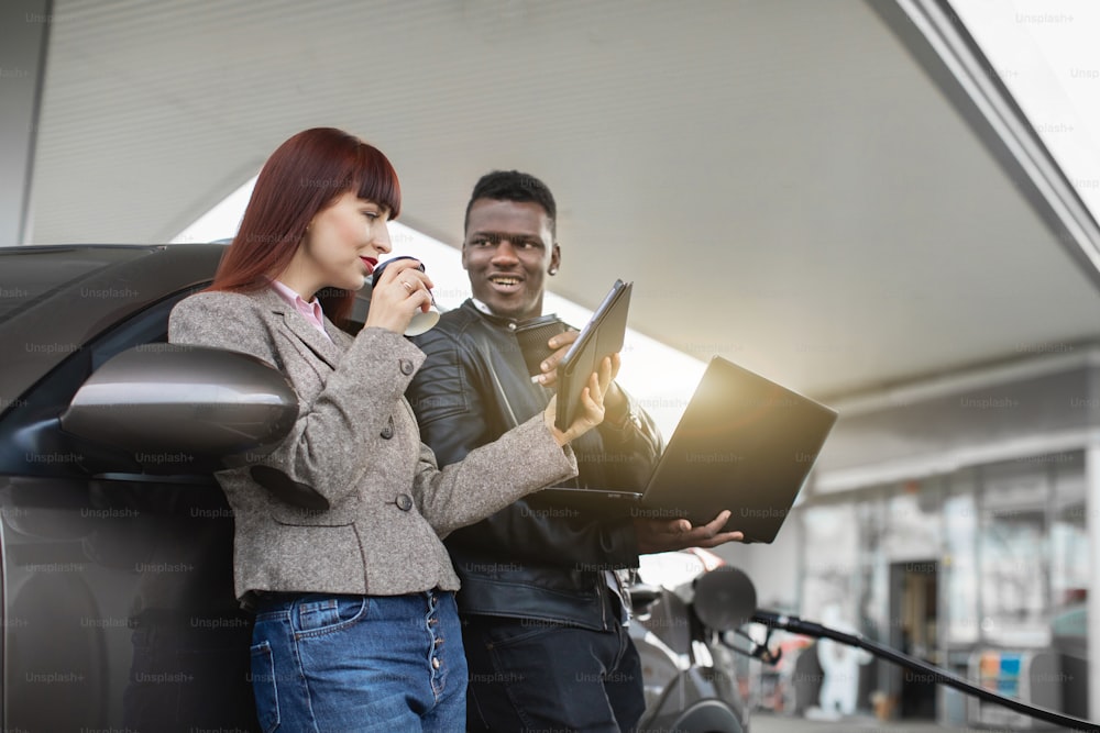 happy multiethnic couple or business colleagues, African man and Caucasian woman holding paper cups with coffee and using tablet and laptop pc, while refueling car at gas station.