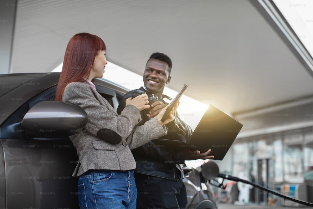 Close up horizontal shot of multiracial laughing couple, leaning on luxury car, talking each other, holding tablet and laptop, drinking coffee to go, refueling the car at gas station.