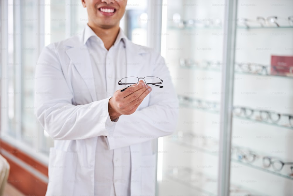 Foto recortada de un optometrista sonriente y contento sosteniendo un par de anteojos en sus manos