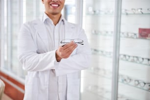 Photo recadrée d’un optométriste souriant et satisfait tenant une paire de lunettes dans ses mains