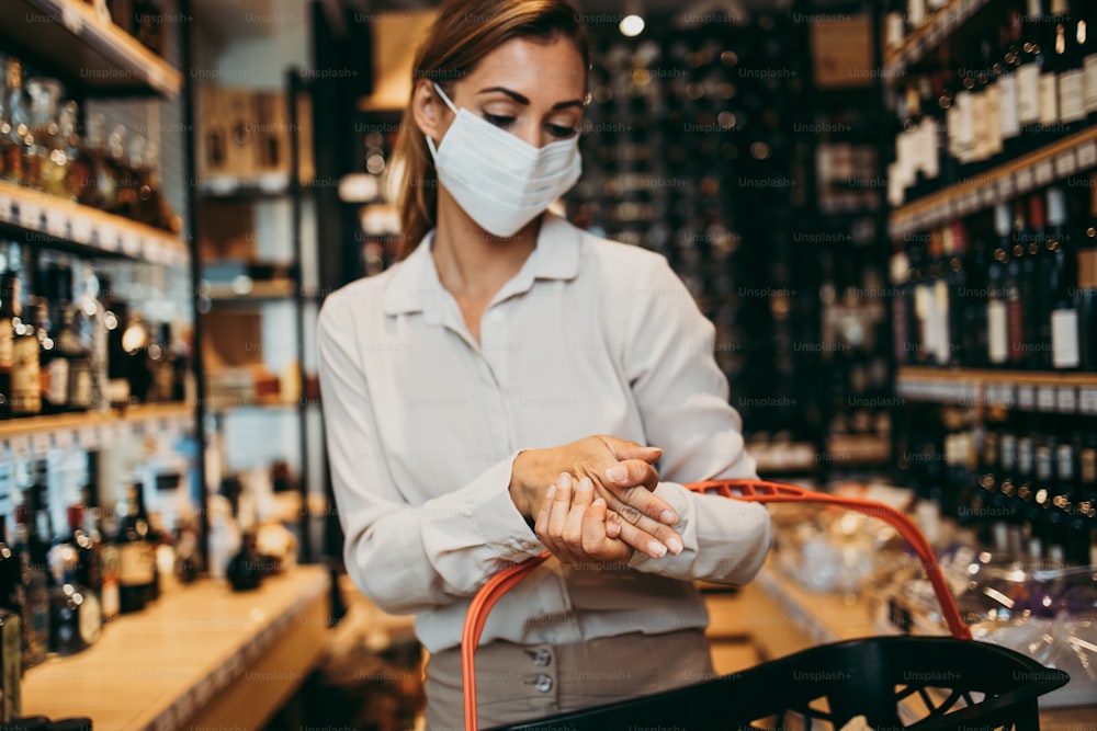 Beautiful young and elegant woman with face protective mask and gloves buying healthy food and drink in a modern supermarket or grocery store. Pandemic or epidemic lifestyle and consumerism concept.