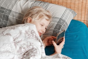 Cute girl lying in bed in morning and texting talking at social media on smartphone cellphone. Child kid typing messaging on mobile phone. Chat talk with friends online on Internet. Video chat.