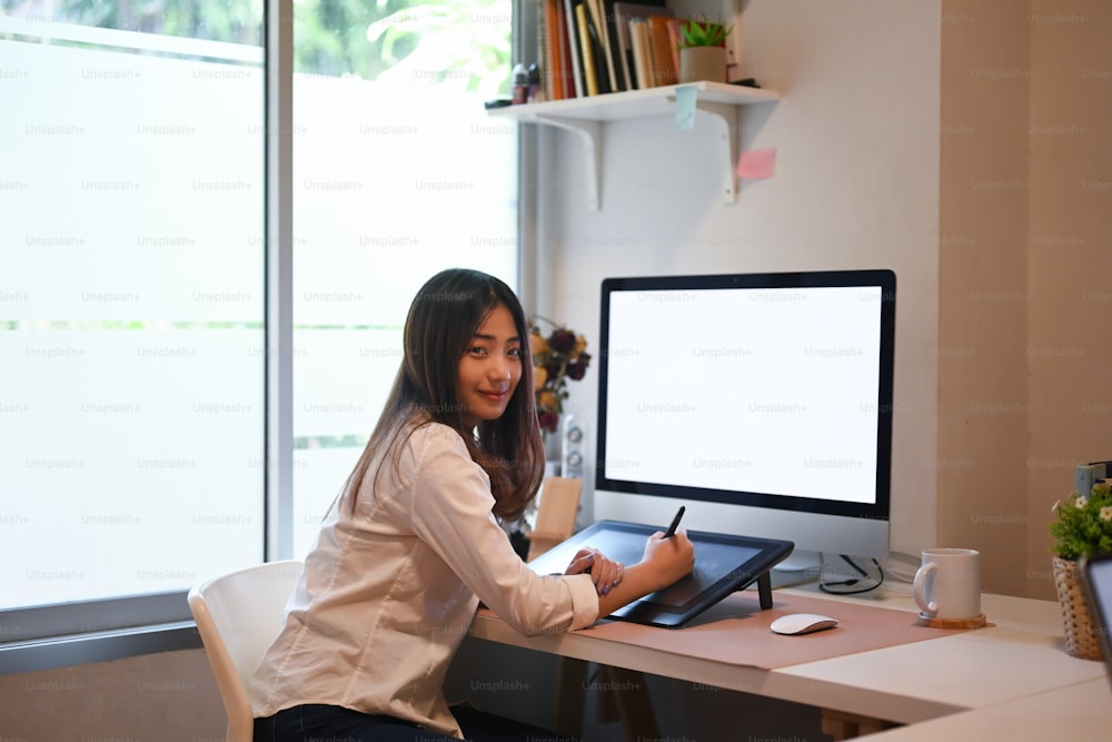 Jovem designer gráfica feminina trabalhando em digitalizador e computador no local de trabalho moderno e olhando para a câmera.