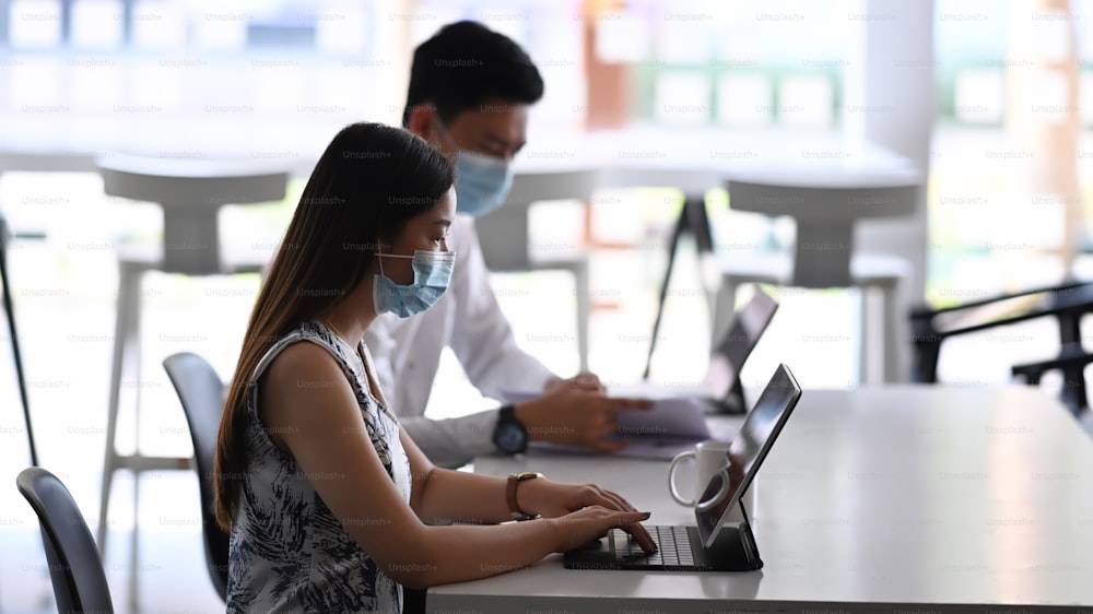 Side view of businesspeople wearing protective mask working with tablet computer and sitting together in modern office.
