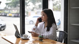 Giovane donna che parla al telefono cellulare e tiene in mano la carta di credito che effettua il pagamento online o lo shopping online mentre è seduta al bar.