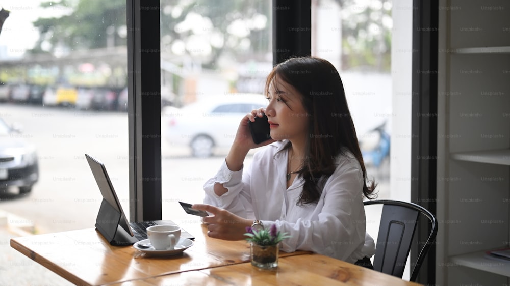 Young female talking on mobile phone and holding credit card making payment online or online shopping while sitting at cafe.