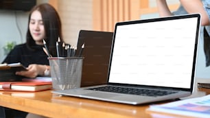 Close up view of laptop computer with blank screen wooden table and young female designer sitting in background.