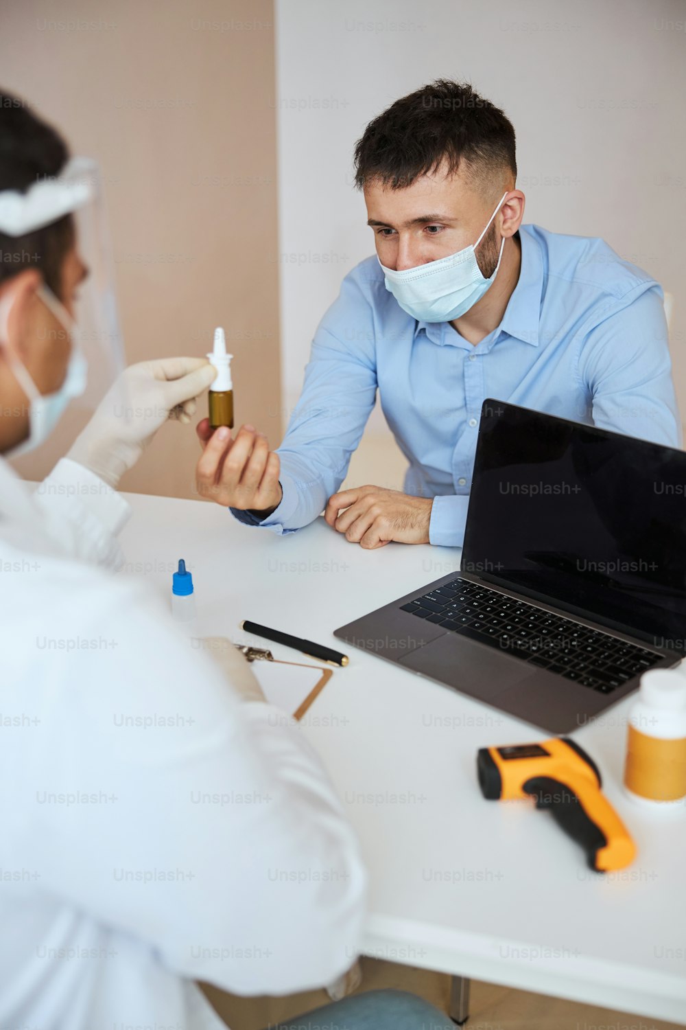 Taillenporträt eines Mannes im blauen Hemd in Medizinmaske, der mit dem Therapeuten spricht, während er eine Kur im Arztkabinett in Betracht zieht