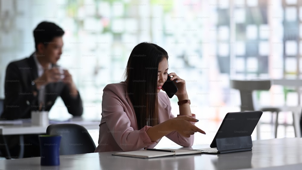 Geschäftsfrau, die ein Geschäftsgespräch auf dem Handy führt und mit einem Tablet-Computer im modernen Büro arbeitet.
