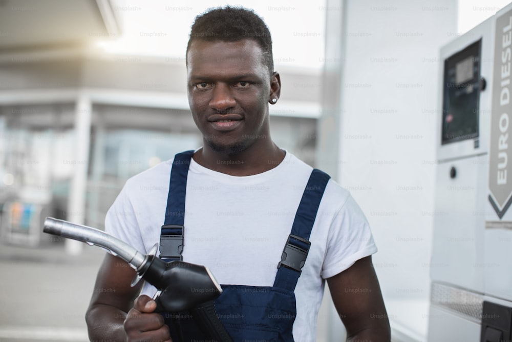 Close up portrait of gas station worker, handsome young African American man, wearing white t-shirt and blue overalls, posing to camera with smile, holding filling gun nozzle.