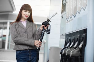 Retrato de primer plano de una atractiva joven caucásica sonriente, sosteniendo una boquilla de gasolina de repostaje y posando ante la cámara en la gasolinera.