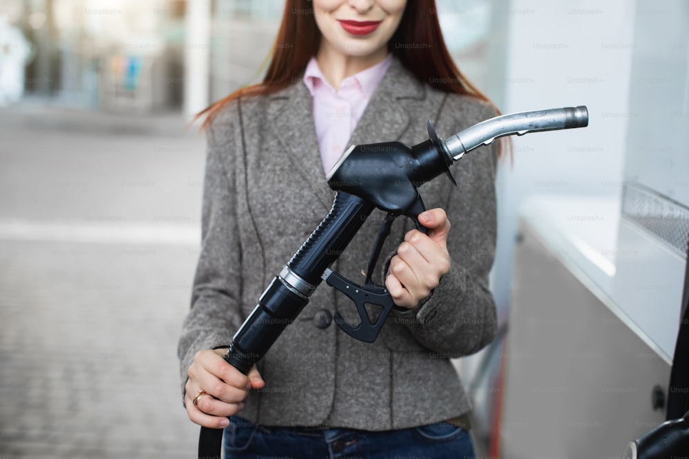 Vista de perto da jovem mulher de negócios caucasiana em traje casual inteligente da moda, segurando a arma do bico de combustível no posto de gasolina pronta para reabastecer o carro.