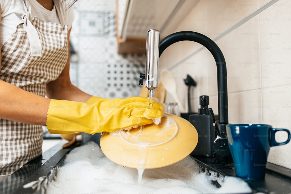 Giovane donna adulta con guanti protettivi gialli che lava i piatti sul lavello della cucina. Routine di igiene domestica e domestica.