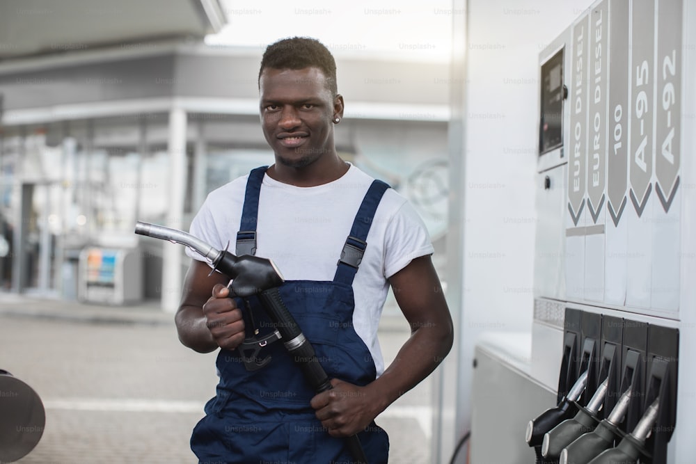 ガソリンスタンドの労働者である作業服を着た若いアフリカ人男性は、車に燃料を補給する準備ができており、充填ガンノズルを持ち、笑顔でカメラを見ています。