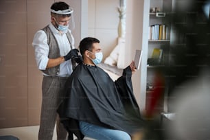 Waist up side view portrait of professional hair stylist in black gloves and medicine mask with comb in hand brushing client hair while visitor looking to yourself in the mirror