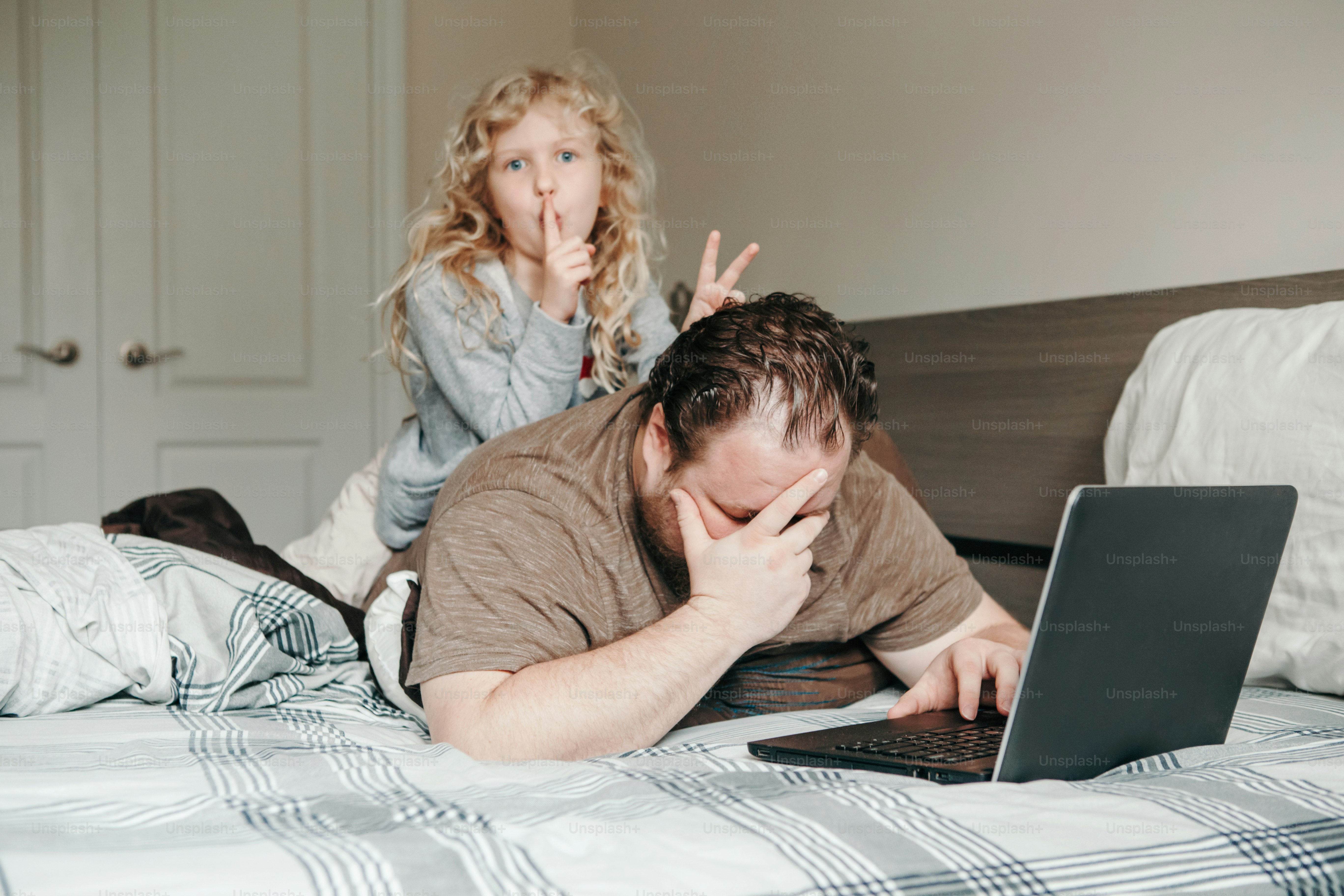 Trabaja desde casa con niños. Padre trabajando en una computadora portátil en el dormitorio con una hija pequeña en su espalda. Momento familiar divertido y sincero. Nueva normalidad durante el confinamiento por la cuarentena por el coronavirus.