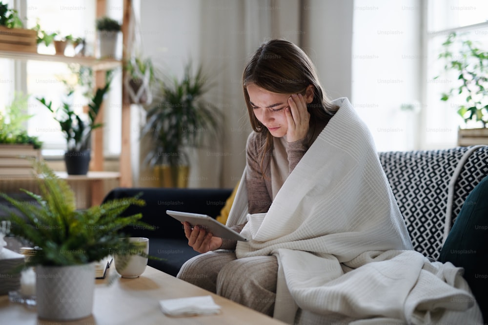 Giovane donna sconvolta e malata avvolta in una coperta sul divano di casa, usando il tablet.