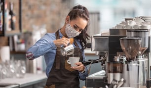 Waitress prepares capuccino in a cafe wearing protective face mask.