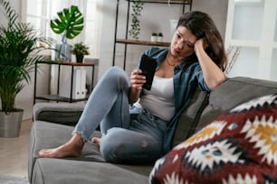 Sad woman sitting on the couch, using the phone. Upset woman waiting for a phone call