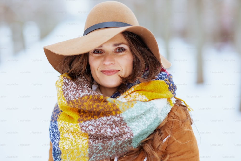 Portrait of smiling stylish middle aged woman in brown hat and scarf outdoors in the city park in winter in sheepskin coat.