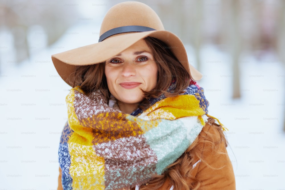 Portrait of smiling stylish middle aged woman in brown hat and scarf outdoors in the city park in winter in sheepskin coat.